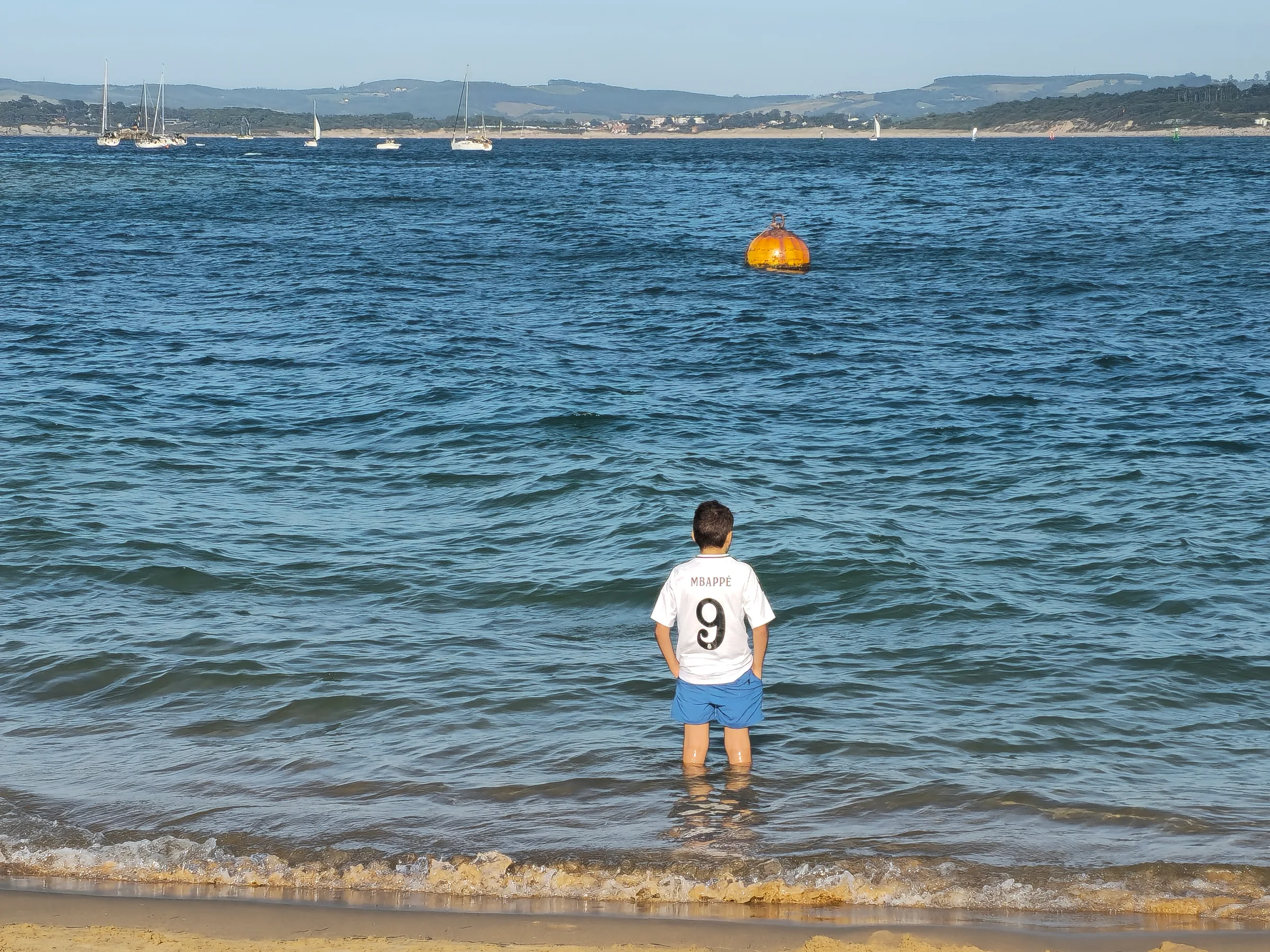 Niño en la orilla de la bahía de Santander en verano con veleros al fondo